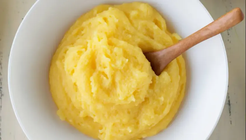 Un tazón blanco lleno de polenta cremosa, con una cuchara de madera. ¿Sabes qué es la polenta? Es un plato reconfortante.