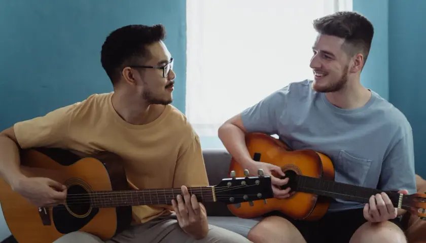 Dwóch mężczyzn gra na gitarach akustycznych. Jeden z nich ma okulary, drugi uśmiecha się.