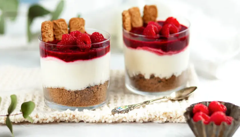 Zwei Gläser mit Schichtdessert: Keksboden, cremige Füllung, Himbeersoße und frische Himbeeren. Perfekte nachtisch ideen für jeden Anlass.