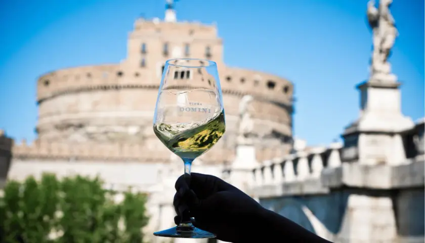Un calice di vino bianco ondeggia con il logo "Vinea Domini" sullo sfondo di Castel Sant'Angelo a Roma.