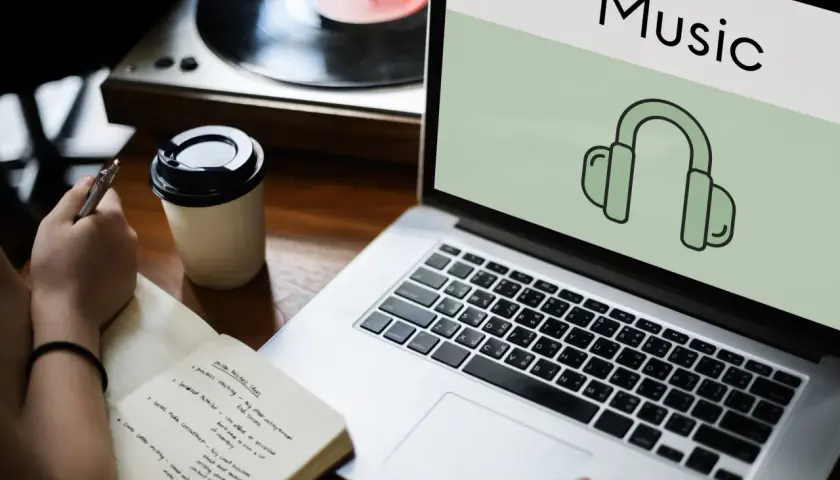 Osoba notuje pomysły na biznes, obok stoi kubek kawy i laptop z ikoną słuchawek, sugerującą aplikację do rozpoznawania muzyki.