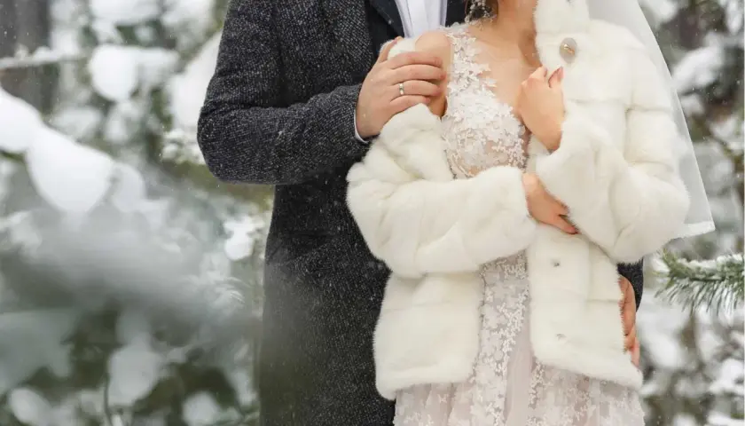 Para młoda w zimowej scenerii. Ona w białym futerku i koronkowej sukni, on w eleganckim płaszczu. Ich ślub zimą wygląda bajkowo.