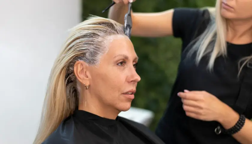 Fryzjerka nakłada farbę na blond włosy klientki. Czy ten kolor włosów postarza? Kobieta ma na sobie czarny fartuch.