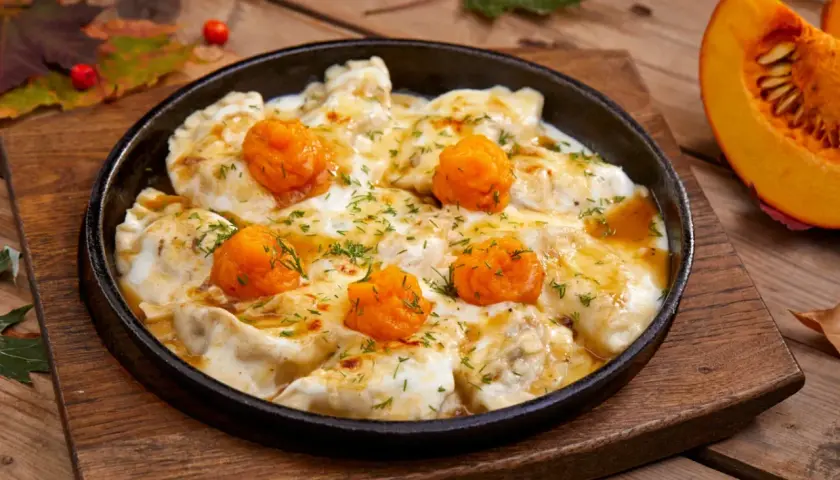 Pierożki z dynią w sosie, posypane koperkiem, podane w żeliwnej patelni na drewnianym blacie. Obok kawałek dyni.