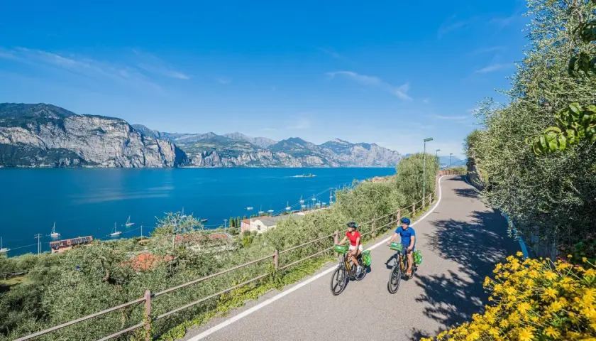 Two people enjoy biking along a scenic road in Italy, with a stunning blue lake and mountains in the background.