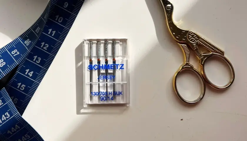 Schmetz Jersey needles, size 90/14, are ideal for stretch fabrics. A blue measuring tape and gold scissors complete the sewing setup.
