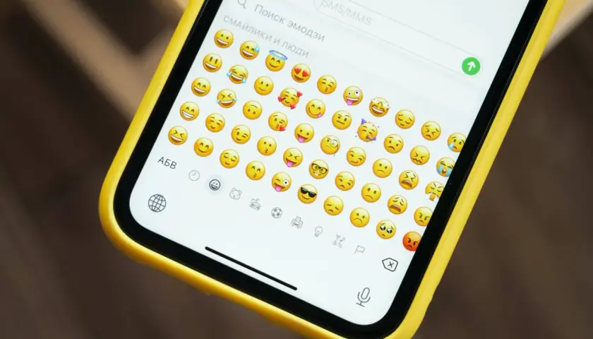 Na ekranie telefonu w żółtej obudowie widać klawiaturę z mnóstwem wesołych i smutnych emotek.