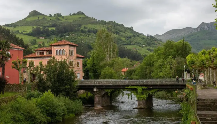 Un puente cruza un río, conectando pueblos bonitos cerca de Liérganes. Edificios rosados y verdes colinas completan la escena.