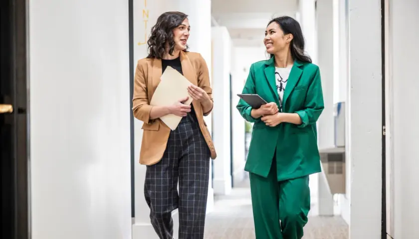 Zwei Frauen in lässiger Business-Kleidung gehen durch einen Flur. Eine trägt einen braunen Blazer und karierte Hose, die andere einen grünen Hosenanzug.