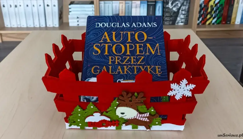 Książka "Autostopem przez galaktykę" Douglasa Adamsa w czerwznym, świątecznym koszyku z reniferem i choinkami.