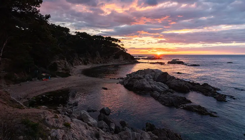 Atardecer en la punta estrella Moraira, con el sol asomando entre rocas y el mar reflejando los tonos rosados del cielo.