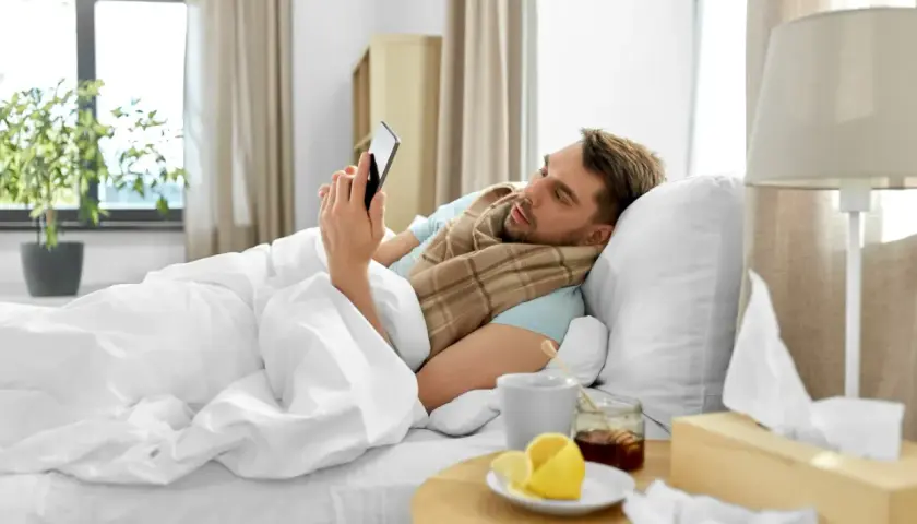 Chory mężczyzna w łóżku, owinięty szalikiem, przegląda telefon. Obok kubek z herbatą, cytryna i miód. Czyżby czekał na zwolnienie lekarskie z powodu siły wyższej, zgodnie z kodeksem pracy?