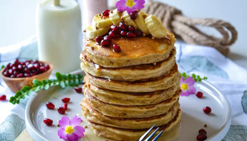 Pile de pancake banane avoine, garnie de fruits rouges, de rondelles de banane et de fleurs roses. Un délice matinal !