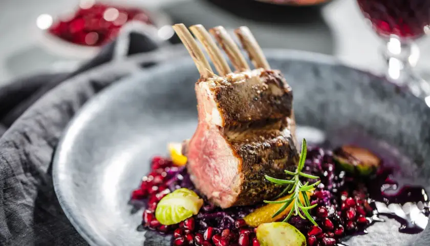 Carré d'agneau parfaitement rôti, servi avec une garniture colorée de grenade, chou rouge et romarin. La cuisson du carré d'agneau est mise en valeur par la présentation.