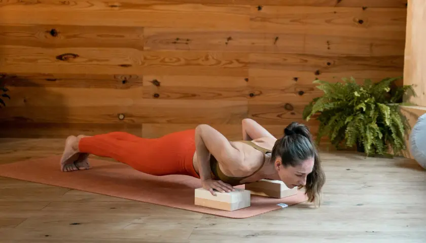 Kobieta w pomarańczowych legginsach wykonuje pozycję chaturanga dandasana, opierając dłonie na drewnianych klockach.