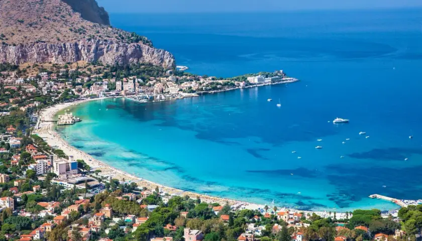 Vista aerea della spiaggia di Palermo, con acque turchesi, case colorate e montagne sullo sfondo.