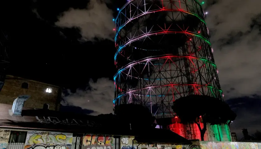 Il gasometro di Roma, illuminato da luci colorate, si staglia contro il cielo notturno. Un muro di graffiti decora l'edificio sottostante.