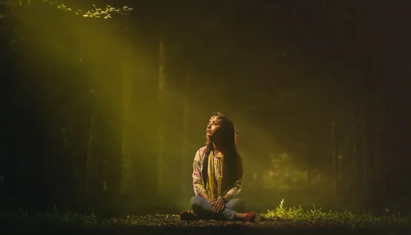 En ung kvinna sitter i skogen, badad i ett gyllene ljus. Hon ser uppåt, som om hon söker svar, kanske om vad gaia betyder.