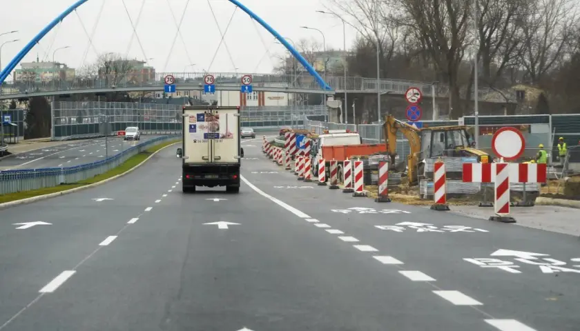 Droga w budowie, z koparką i barierami. W tle niebieski most i **aleja** z ruchem samochodowym.