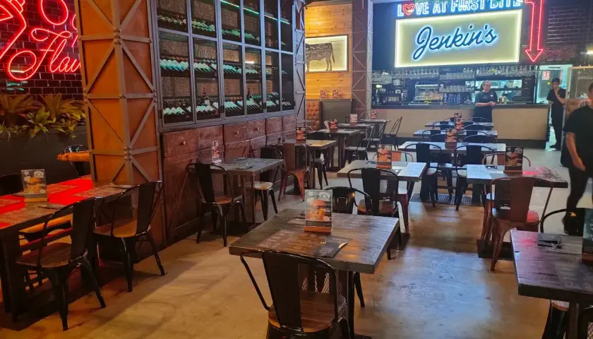 Interior de Jenkins Valencia, un restaurante con mesas de madera, sillas metálicas y un letrero de neón que dice "Love at First Bite Jenkins".