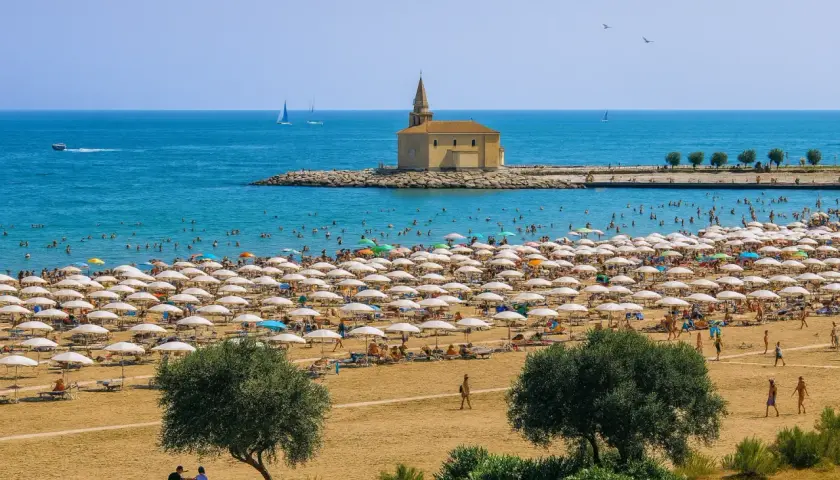 Vue aérienne d'une plage bondée à Caorle, avec des rangées de parasols blancs, des baigneurs dans la mer bleue et une petite église sur un îlot rocheux.