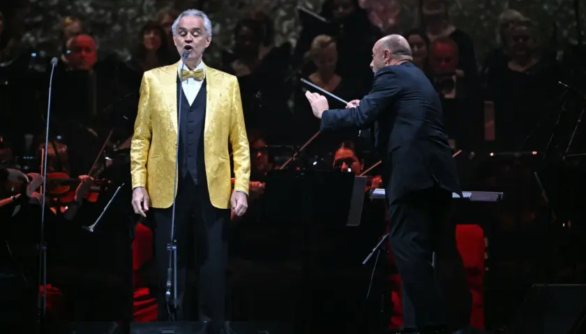 Andrea Bocelli w złotej marynarce śpiewa na scenie, dyrygent prowadzi orkiestrę. Koncert zapowiada się na niezapomniane przeżycie, jakiego można spodziewać się po Andrea Bocelli koncert 2026.