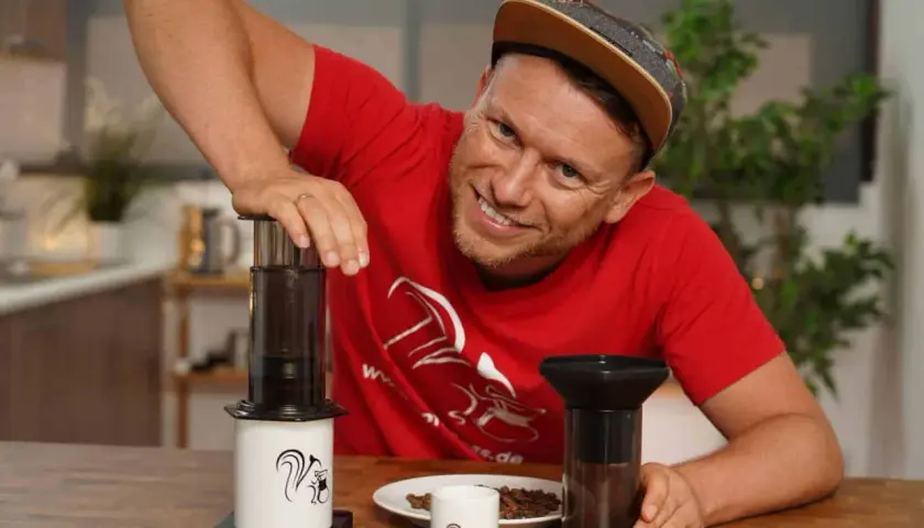Mann mit rotem T-Shirt zeigt die Aeropress Zubereitung mit zwei Geräten und Kaffeebohnen.