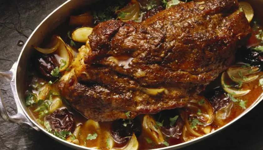 Rouelle de porc rôtie dans un plat avec des oignons caramélisés, des pruneaux et du persil frais.