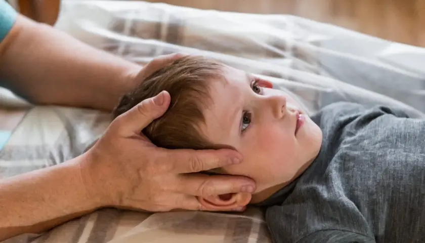 Chłopiec z obniżonym napięciem mięśniowym leży na plecach, a terapeuta delikatnie dotyka jego głowy.