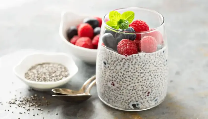Pudding chia z owocami leśnymi, pokazujący jak przygotować nasiona chia. Obok miseczka z nasionami i łyżeczki.