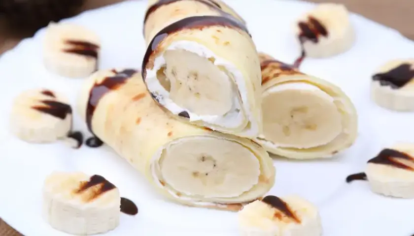 Rolowane naleśniki z bananem i bitą śmietaną, polane czekoladą. Obok ułożone plasterki banana z polewą.