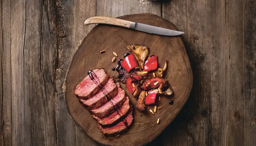 Saftige Tagliata di Manzo mit gebratenen Pilzen und Paprika auf einem rustikalen Holzbrett, garniert mit Pinienkernen und Balsamico.