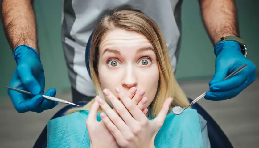 Occhi sbarrati e mani sulla bocca: la paura del dentista è palpabile mentre il dentista si avvicina con gli strumenti.