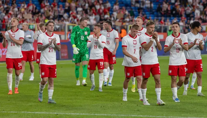Piłkarze reprezentacji Polski w białych strojach z orzełkiem na piersi, klaskający na murawie. Ich pozycja w tabeli mistrzów Polski w piłce nożnej jest nieznana.