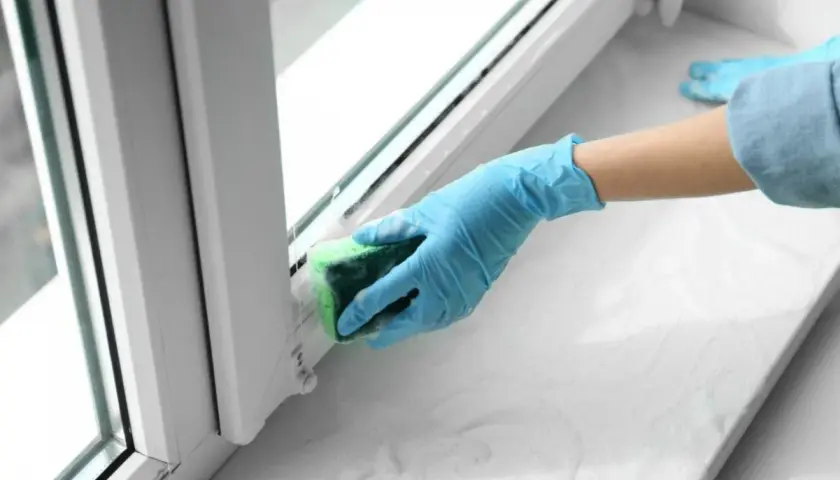 Ręka w niebieskiej rękawiczce czyści parapet gąbką. Dowiedz się, jak usunąć klej po taśmie z drzwi i innych powierzchni.