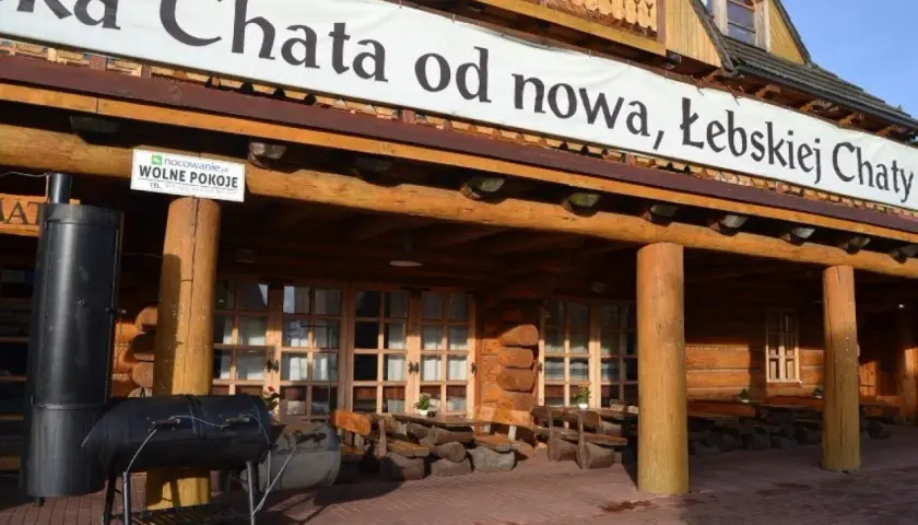 Drewniana, łebska chata z banerem "od nowa, Łebskiej Chaty". Przed wejściem grill i stoliki.