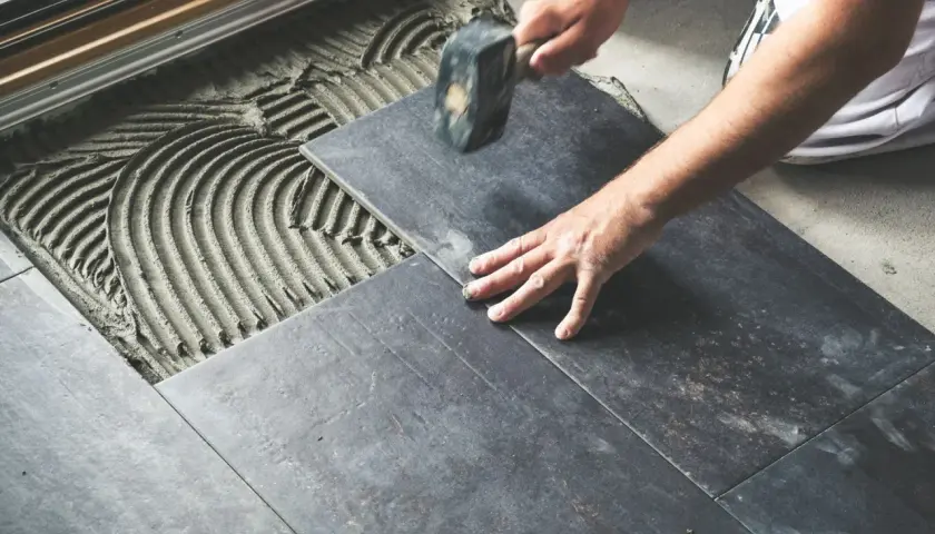 Mano che posa una piastrella scura su cemento fresco, con un martello per un posizionamento perfetto e rettificato.
