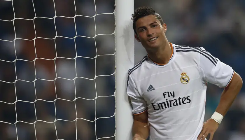 Uśmiechnięty Cristiano Ronaldo opiera się o słupek bramki. Jego sylwetka i sportowa forma, choć nie widać tu konkretnych danych o Ronaldo Wzrost i Waga, świadczą o jego atletyzmie.