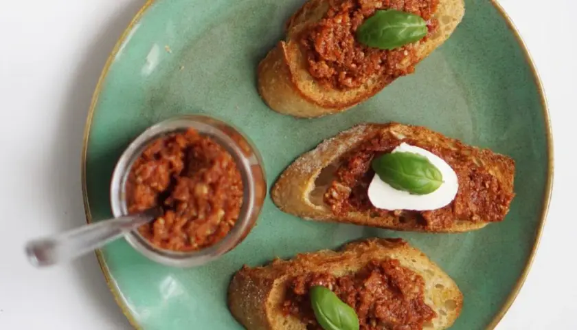Chrupiące grzanki z aromatycznym pesto pomidorowym i listkiem bazylii. Obok słoiczek z pastą.