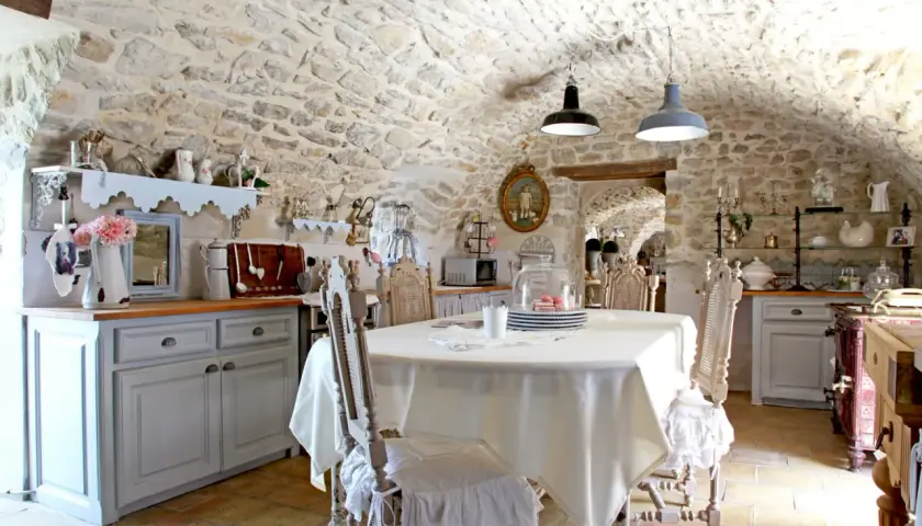 Cuisine rustique au style shabby chic, avec murs en pierre, table dressée et chaises anciennes.