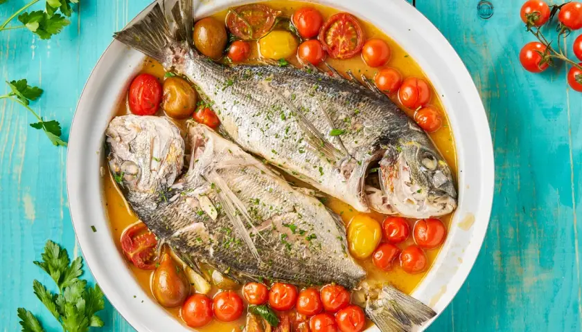Orata all'acqua pazza, due pesci interi cotti in un brodo saporito con pomodorini e erbe aromatiche, serviti in un piatto bianco su un tavolo di legno blu.