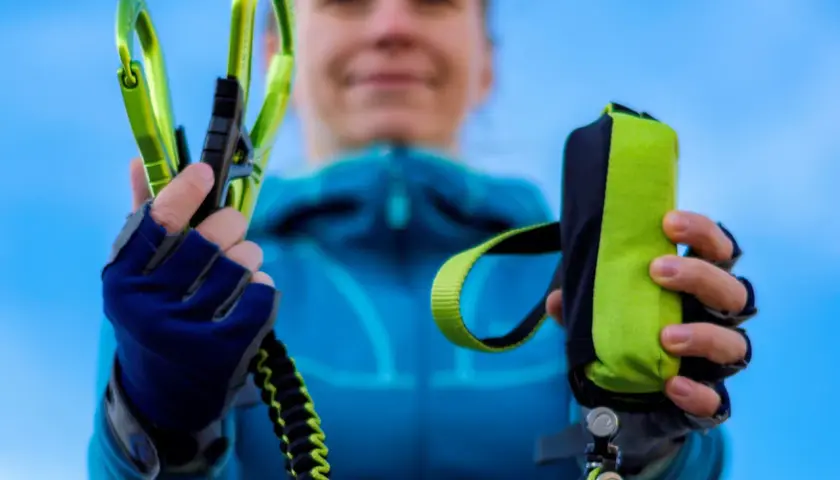 Ręce w rękawiczkach trzymają zielone karabinki i woreczek na via ferrata sprzęt.