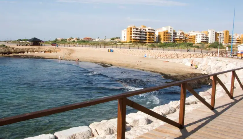 Paseo de madera junto al mar en la cala Piteras. Edificios modernos y playa de arena dorada.