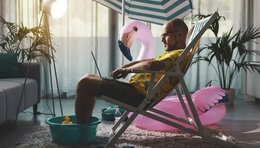 Mężczyzna w hawajskiej koszuli pracuje na laptopie, siedząc w leżaku z nogami w misce. W tle dmuchany flaming i parasol plażowy. Czas wykorzystać zaległy urlop!