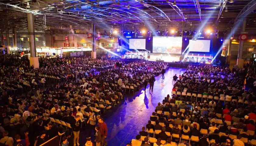 Gran multitud en un evento de convencion, sentada en sillas frente a pantallas gigantes con luces de escenario.