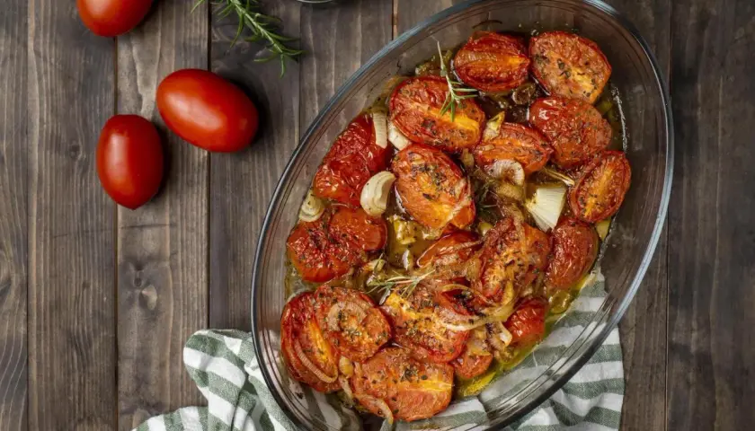 Pomodori gratinati al forno, dorati e succosi, con cipolle e rosmarino in una teglia di vetro su un tavolo di legno.