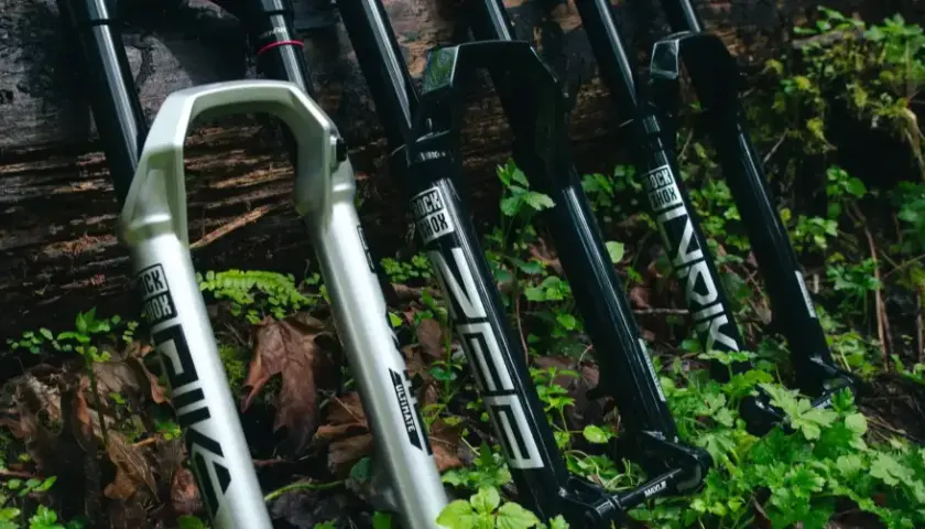 A silver RockShox Pike and black RockShox Lyrik forks lean against a log, ready for trail adventures.