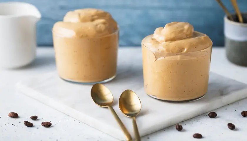 Due bicchieri di cremosa schiuma di caffè, perfetti per una ricetta crema di caffè senza panna. Due cucchiaini dorati e chicchi di caffè completano la scena.