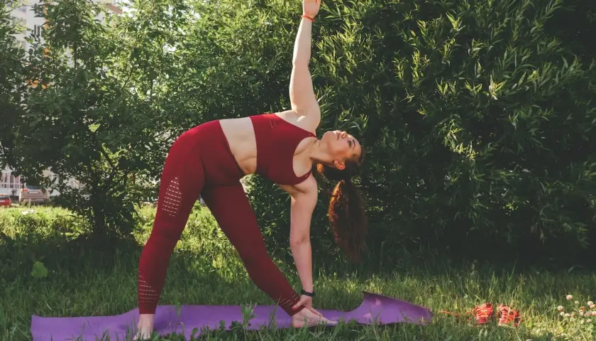 Kobieta w czerwonym stroju wykonuje pozycję Trójkąta podczas praktyki jogi ashtanga na trawie, otoczona zielenią.