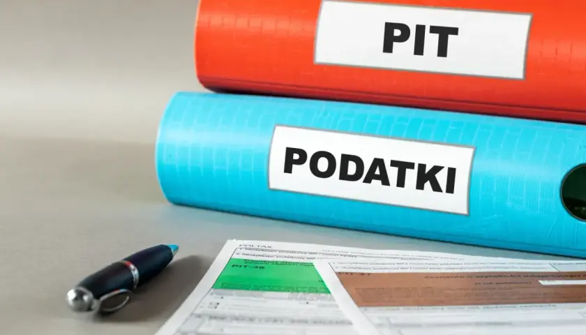 Dwa segregatory: czerwony z napisem "PIT" i niebieski z napisem "PODATKI". Obok formularze podatkowe i długopis. Ulga dla młodych w zasięgu ręki.